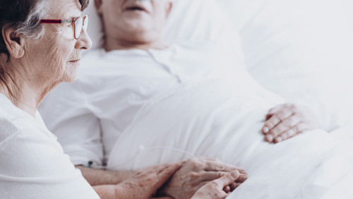 Vrouwelijke mantelzorger zit naast zieke man die in bed ligt in de laatste fase Vrouwelijke mantelzorger zit naast zieke man die in bed ligt in de laatste fase