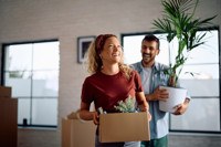 Een jong stel staat in hun nieuwe woning, de vrouw draagt verhuisdozen en de man een plant, ze lachen Een jong stel staat in hun nieuwe woning, de vrouw draagt verhuisdozen en de man een plant, ze lachen