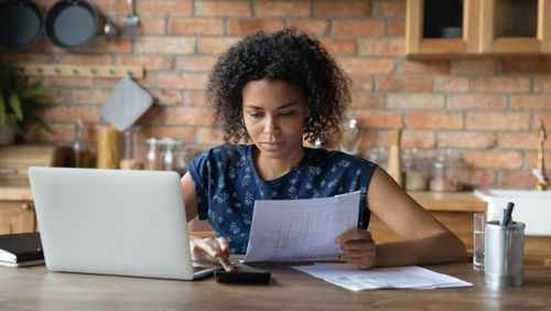 Een jonge vrouw zit in de keuken achter de laptop en maakt overzicht van haar financien Een jonge vrouw zit in de keuken achter de laptop en maakt overzicht van haar financien